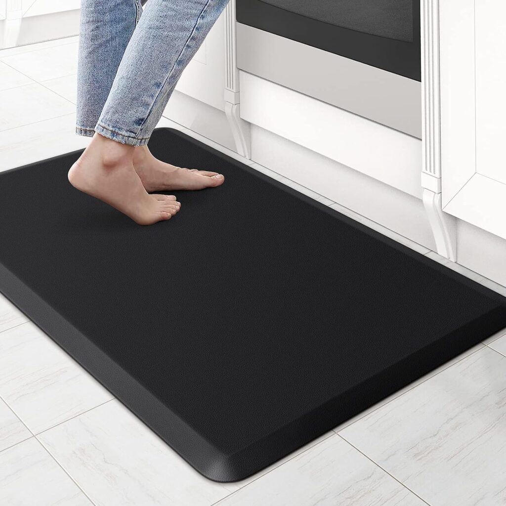 Black anti-fatigue mat placed on a kitchen floor, with a person standing on it barefoot. The mat has a thick, cushioned design to provide comfort and support, reducing strain on the feet and legs during prolonged standing. The surrounding kitchen features white cabinetry and a tiled floor.