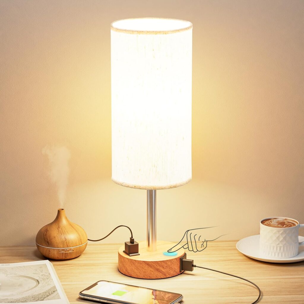 Bedside table lamp with a cylindrical fabric shade, emitting a warm light. The lamp features a wooden base with a touch control button for adjusting brightness. It also includes two USB charging ports, shown in use with a phone and a diffuser. The setup is on a wooden table alongside a cup of coffee and an open book.