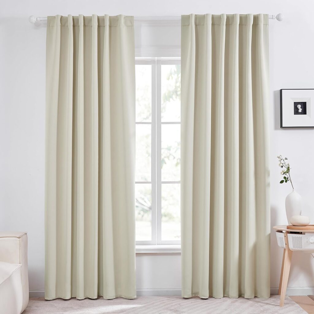 A bright and airy living room featuring cream-colored thermal insulated curtains from Deconovo. The curtains are neatly hung on a white curtain rod, framing a large window that lets in natural light. The room includes a small side table with a white vase and a few decorative items, enhancing the cozy and inviting atmosphere.