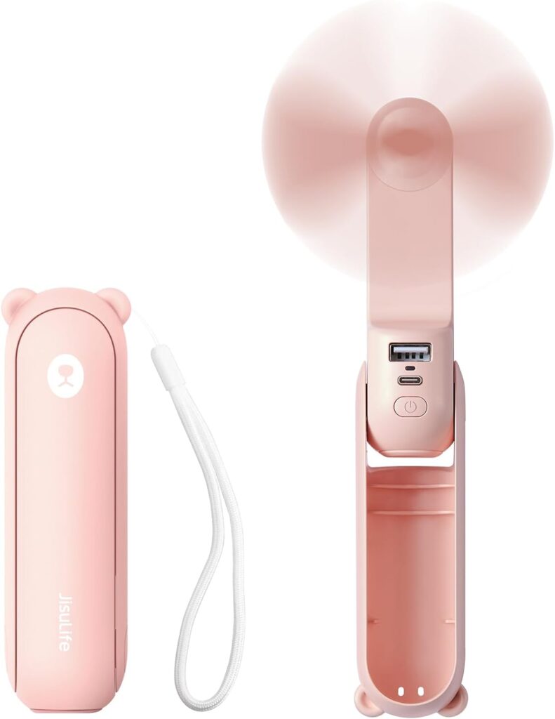 Pink handheld mini fan with a compact design, featuring a bear ear motif on the cover. The fan includes a wrist strap for easy carrying and displays a USB port, a power button, and charging port on the back. The fan blades are shown in motion, indicating it is turned on.